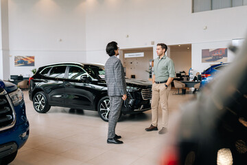 Remote view of doubtful male customer buying car in showroom talking to sales agent in business suit looking at auto indoors in spacious dealership. Concept of choosing and buying auto at showroom.