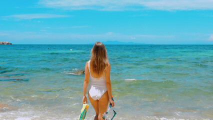 Sensual young woman holds snorkel mask and fins, ready for underwater snorkeling. Thailand travel, summer vibes. Concept of beach vacation and water adventure.