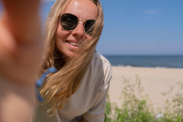 Young woman in sunglasses with shell bracelets looking into camera. Influencer Using mobile phone for video call selfie waving on the beach seacoast. Holiday vacation advertisement concept. Social