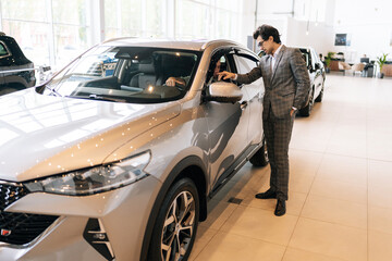 Wide shot of successful customer male talking to sales representative in motor showroom sitting inside brand new car holding steering wheel. Concept of buying new auto in dealership office.