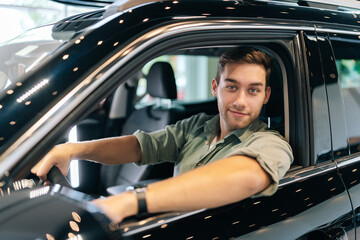 Fototapeta na wymiar Portrait of happy confident customer male client putting hand on steering wheel, looking at camera while trying new automobile at showroom. Satisfied young man client doing drive test of new car
