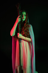 Fototapeta premium portrait of beautiful brunette woman wearing a gown with purple fantasy cloak holding a wooden wizard staff, isolated on dark studio background with cinematic colourful lighting. 