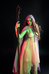 portrait of beautiful brunette woman wearing  a gown with purple fantasy cloak holding a wooden wizard staff, isolated on dark studio background with cinematic colourful lighting.
