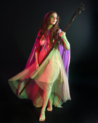 Obraz na płótnie Canvas portrait of beautiful brunette woman wearing a gown with purple fantasy cloak holding a wooden wizard staff, isolated on dark studio background with cinematic colourful lighting. 