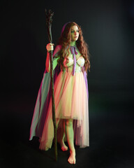 Obraz na płótnie Canvas portrait of beautiful brunette woman wearing a gown with purple fantasy cloak holding a wooden wizard staff, isolated on dark studio background with cinematic colourful lighting. 