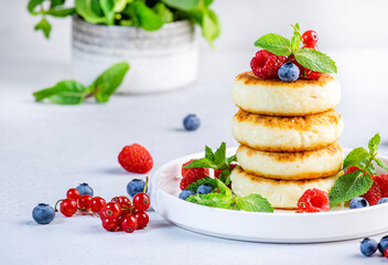 Thick pancakes with cottage cheese with blueberries, raspberries and red currants, decorated with mint on white plate, delicious breakfast. Gray table background