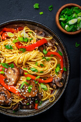 Stir fry noodles with vegetables: red paprika, champignons, green onion and sesame seeds in ceramic bowl. Black table background, top view