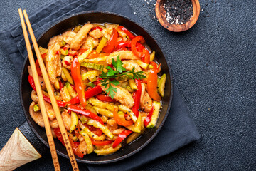 Asian cuisine dish, stir fry chicken, paprika and zucchini, ginger, garlic with sesame and soy sauce in a frying pan. Black table background, top view