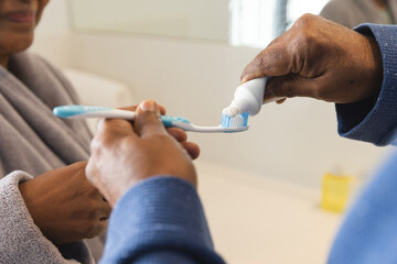Senior biracial couple in bathrobes squeezing toothpaste on toothbrush in bathroom at home