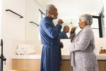 Happy senior biracial couple wearing bathrobes and brushing teeth in bathroom at home - Powered by Adobe