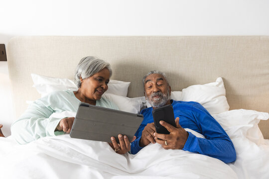Happy senior biracial couple lying in bed and using tablet and smartphone at home