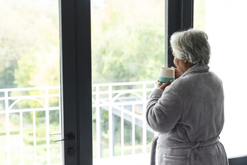 Thoughtful senior biracial woman wearing bathrobe and drinking coffee by window at home