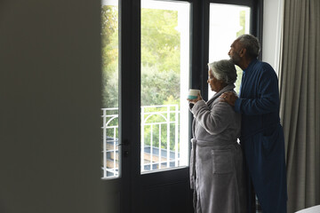 Thoughtful senior biracial couple in bathrobes drinking coffee by window at home, copy space