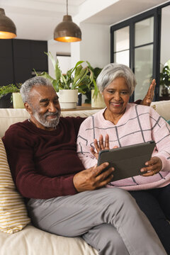 Happy Senior Biracial Couple Sitting On Couch And Having Tablet Video Call At Home