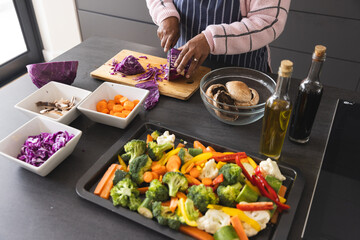 Senior biracial woman wearing apron and cutting cabbage on table with vegetables in kitchen at home