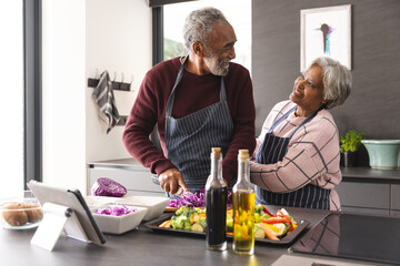 Happy senior biracial couple wearing aprons preparing vegetables in kitchen at home