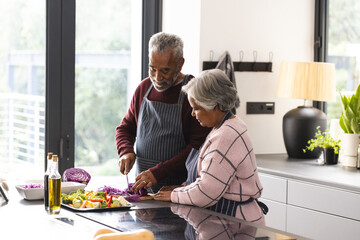 Happy senior biracial couple wearing aprons preparing vegetables in kitchen at home - Powered by Adobe