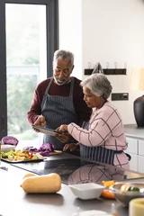 Foto auf Acrylglas Kochen Happy senior biracial couple using tablet preparing vegetables in kitchen at home, copy space