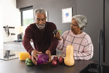 Happy senior biracial couple unpacking bags of vegetables in kitchen at home