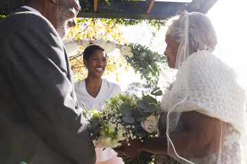 Happy biracial female marriage officiant with senior couple during wedding ceremony in sunny garden