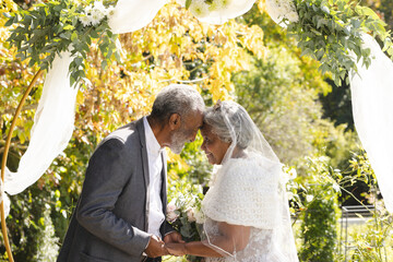 Happy senior biracial bride and groom during wedding ceremony in sunny garden