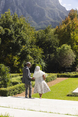 Fototapeta premium Happy senior biracial bride and groom walking in garden at sunny outdoor wedding ceremony