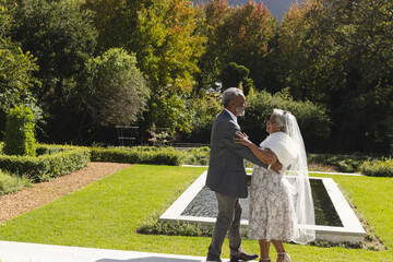 Happy senior biracial bride and groom dancing in garden at sunny wedding ceremony, copy space