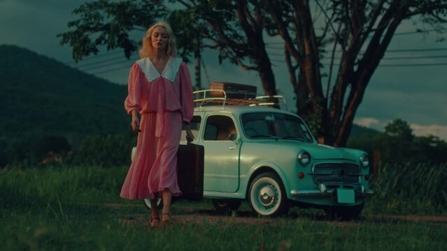 A blonde in a vintage dress picks up her suitcases and walks away with her retro car in the background
