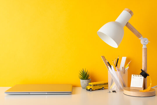 Back-to-school essentials on display. Side view photo of stationery holder, small calendar, graduate hat, flowerpot, laptop, lamp, school bus toy, arranged neatly on a desk against yellow wall