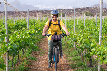 Happy senior woman with helmet riding outdoors on electric bike among vineyards. Sporty elderly...