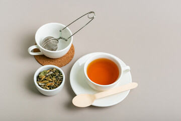 Cup of hot tasty green tea, strainer for tea in empty white mug and herbal tea leaves in ceramic small bowl on light background.