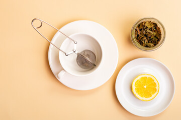 Obraz na płótnie Canvas Green tea leaves, white ceramic cup for tea, tea strainer and lemon on small plate from above on light beige background in rustic style.