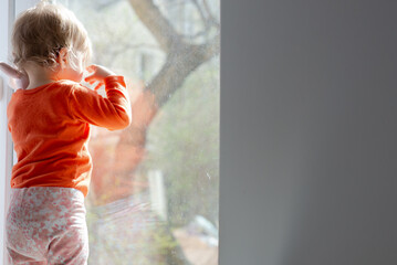 baby girl standing in front of the window glass at sunny day and having a fun with the sunbeams,...