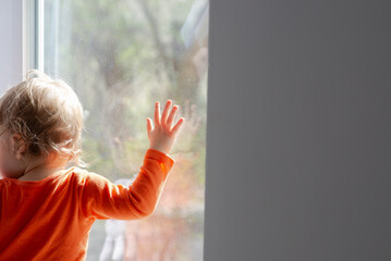 baby girl standing in front of the window and touch glass at sunny day, having fun with the sunbeams, natural dirty home glass - obrazy, fototapety, plakaty