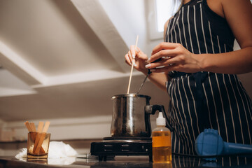 Woman adding essential oil into pot with melted wax at kitchen counter, closeup. Making homemade candles