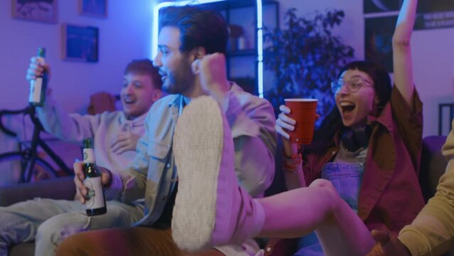 Medium shot of group of young multiethnic friends in casual outfits sitting on couch at home, watching football match on TV, cheering anxiously, whooping at goal and clinking beer bottles and glasses