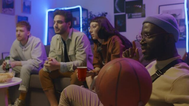 Medium shot of Caucasian girl and guy and Middle Eastern male friend sitting on couch at home, watching sport game on TV, pointing at screen and cheering, and black man fiddling with basketball