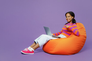 Full body little kid teen IT girl 15-16 years old wear striped orange sweatshirt sit in bag chair hold use work on laptop pc computer isolated on plain purple background. Childhood lifestyle concept.