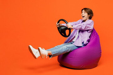 Full body amazed young woman she wear purple shirt white t-shirt casual clothes sit in bag chair hold steering wheel driving car isolated on plain orange background studio portrait. Lifestyle concept.
