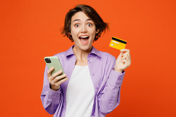 Young caucasian woman wear purple shirt white t-shirt casual clothes using mobile cell phone hold credit bank card do online shopping order delivery booking tour isolated on plain orange background - obrazy, fototapety, plakaty
