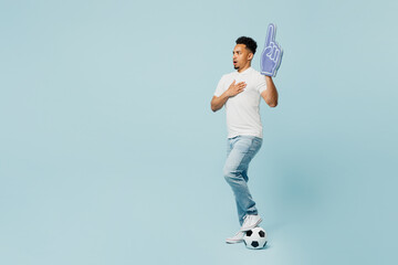 Full body young man fan in t-shirt foam 1 fan glove finger up sing national country anthem cheer up support football sport team hold soccer ball watch tv live stream isolated on plain blue background.