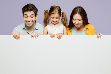Young parents mom dad with child kid daughter girl 6 years old wearing blue yellow casual clothes hold look at big white empty poster billboard isolated on plain purple background. Family day concept.