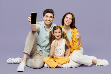 Full body young parents mom dad with child kid daughter girl 6 years old wear yellow casual clothes sit hold use blaank screen mobile cell phone isolated on plain purple background Family day concept