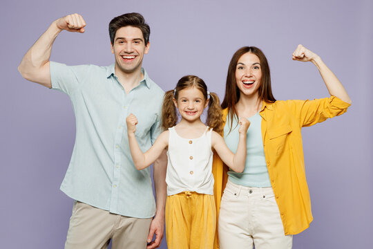 Young Happy Parents Mom Dad With Child Kid Daughter Girl 6 Years Old In Blue Yellow Casual Clothes Show Hand Biceps Muscles Demonstrating Power Isolated On Plain Purple Background. Family Day Concept.