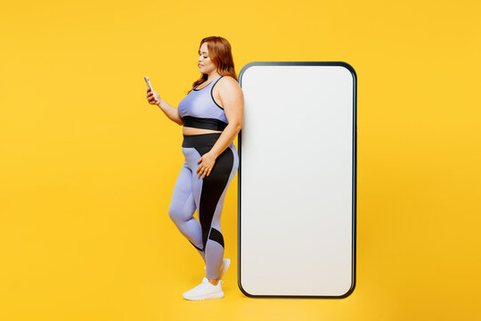 Full body young plus size big fat fit woman wear blue top warm up train big huge black screen mobile cell phone use smartphone isolated on plain yellow background studio home gym Workout sport concept