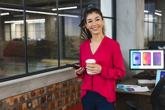 Portrait of biracial female designer holding coffee cup and smartphone smiling at office - Powered by Adobe