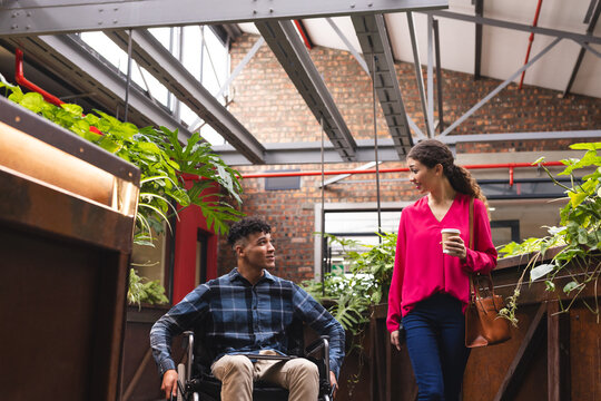 Happy diverse female and male colleague in wheelchair talking in office corridor