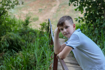 The boy stands on the bridge in nature and looks at the camera with a smile. Space for text.