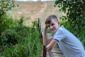 The boy stands on the bridge in nature and looks at the camera with a smile. Space for text.
