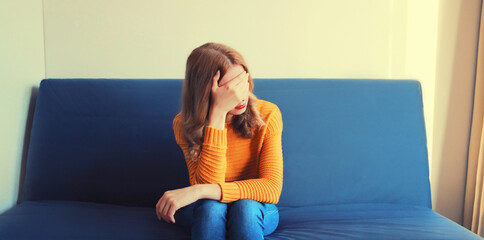 Upset sad crying woman covering her eyes with her hands while experiencing mental or physical suffering on the couch at home